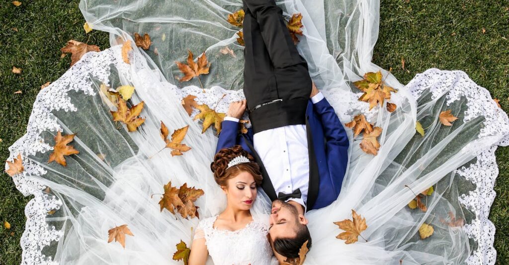 Bride and groom lying together on a lawn surrounded by autumn leaves, captured from above.