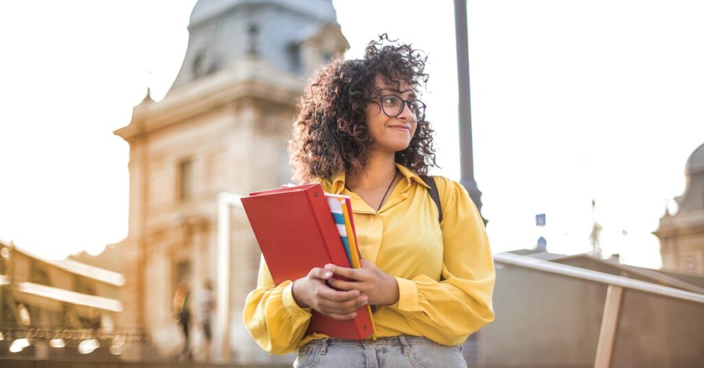 Smiling female student with curly hair and glasses holding books on campus steps.