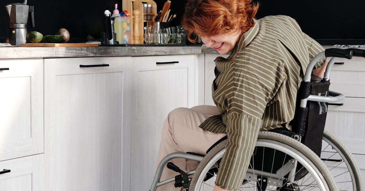 A woman in a wheelchair smiles while petting a tabby cat in a modern kitchen.