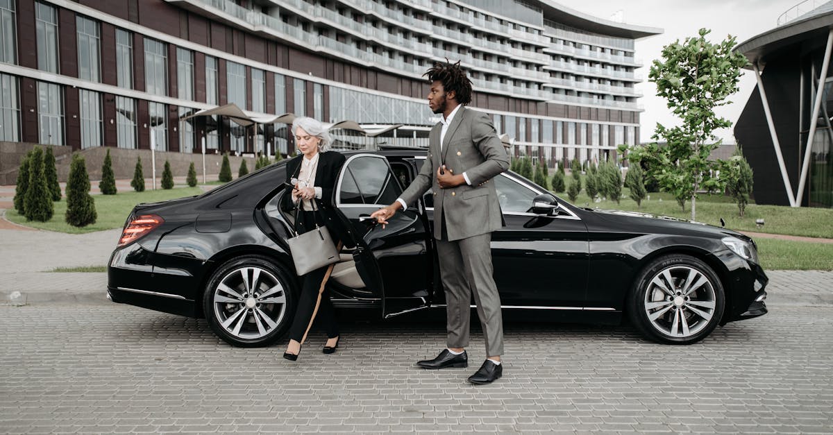 A professional chauffeur assists a businesswoman exiting a luxury car in front of a modern building.